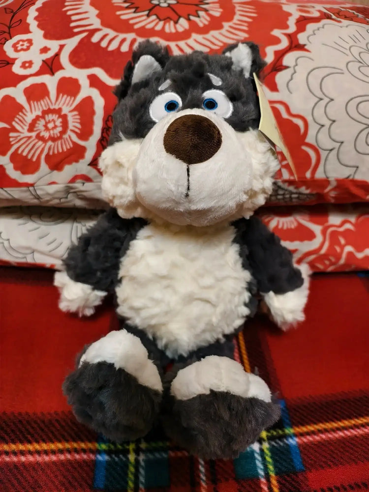 Peluche Loupio le loup assis sur un canapé rouge avec coussin fleuri, fourrure grise et blanche bouclée, grands yeux bleus expressifs.