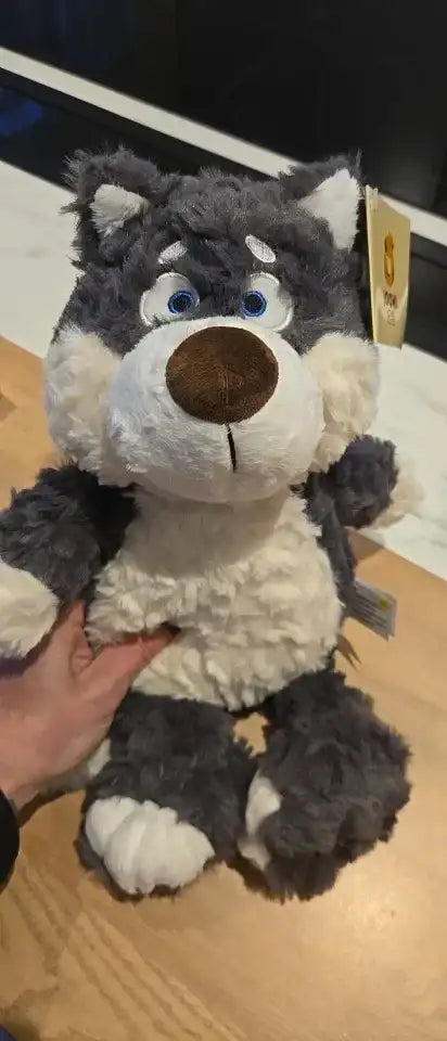 Peluche Loupio le loup tenu à la main, étiquette produit visible, fourrure grise bouclée et ventre crème, grands yeux bleus expressifs.