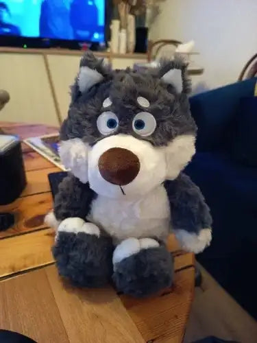 Peluche Loupio le loup posé sur une table en bois dans un salon, fourrure grise et blanche, yeux bleus, ambiance intérieure chaleureuse.