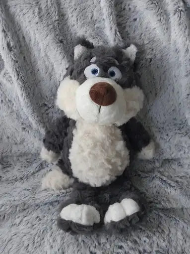 Peluche Loupio le loup assis sur un tapis gris doux, fourrure bouclée grise et blanche, posture détendue, regard expressif.